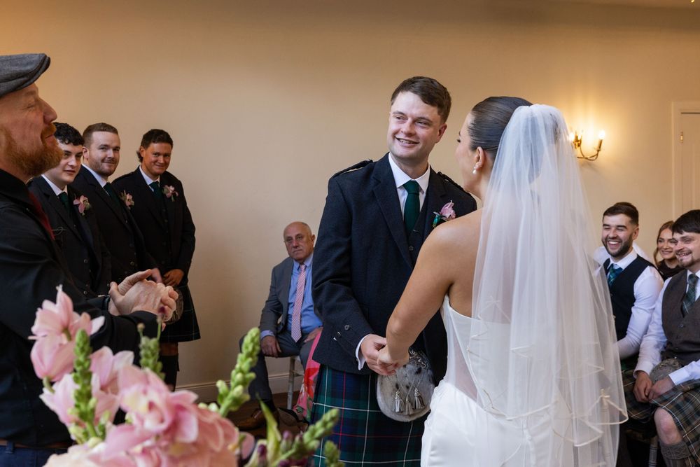 Groom in Scottish kilt smiling at bride in white veil during wedding ceremony with guests in background.