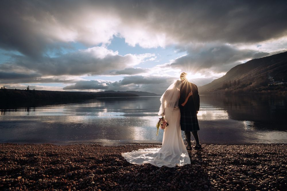 Bride and groom stand by a serene Scottish loch at sunset, with dramatic clouds and misty mountains in the background.