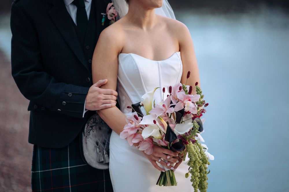 Bride in white strapless gown holds pink lily bouquet beside groom in Scottish tartan kilt near waterfront.
