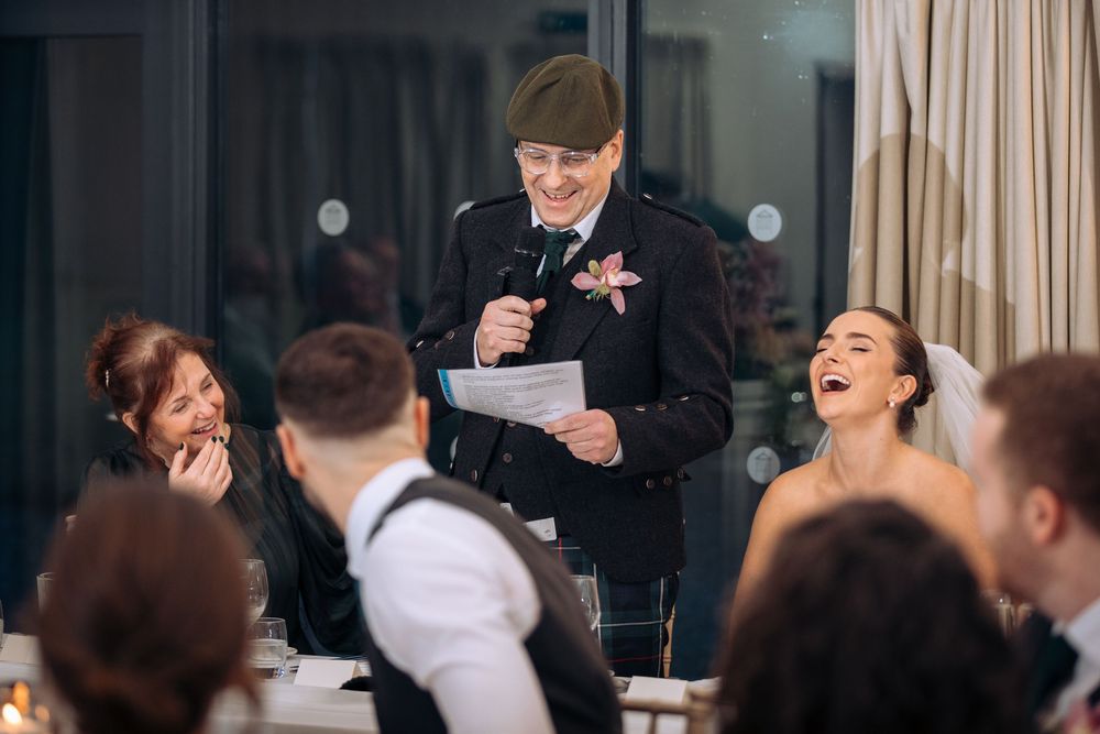 Elderly man in Scottish attire gives a humorous wedding speech as bride laughs joyfully at reception dinner.