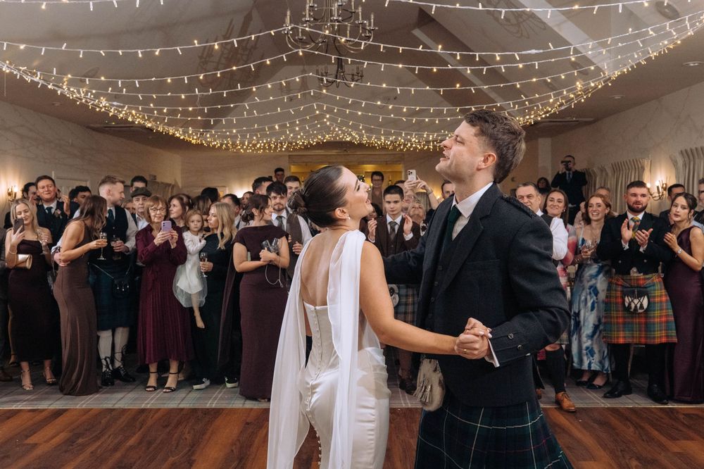 Bride and groom share first dance at Scottish wedding reception with fairy lights and guests in formal attire.