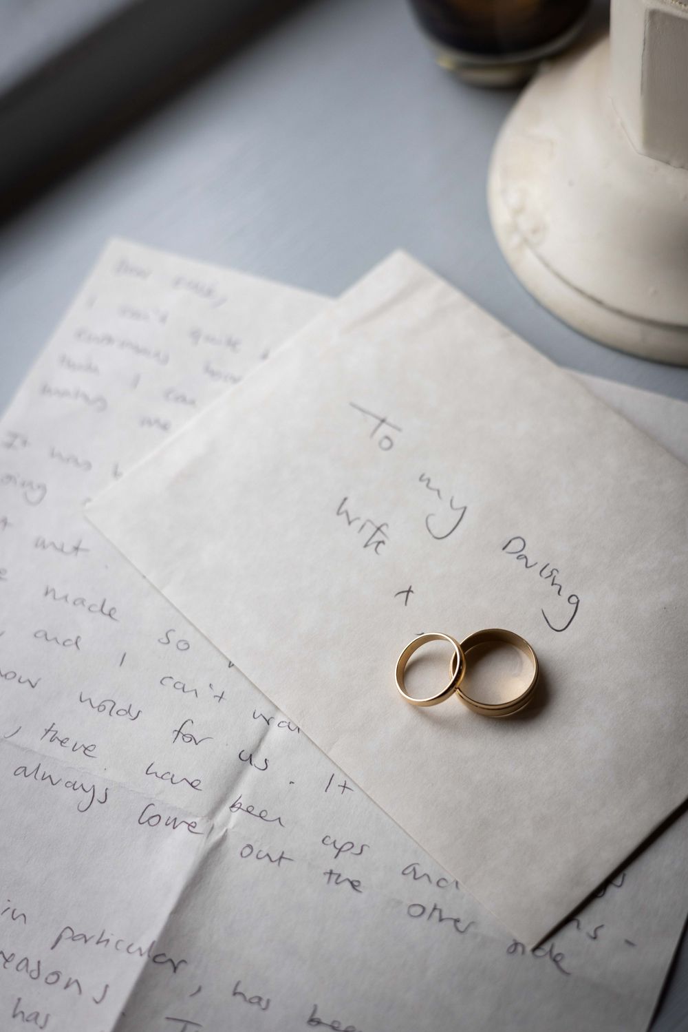 Two gold wedding rings rest on handwritten love letters addressed to my darling wife on a light gray surface.