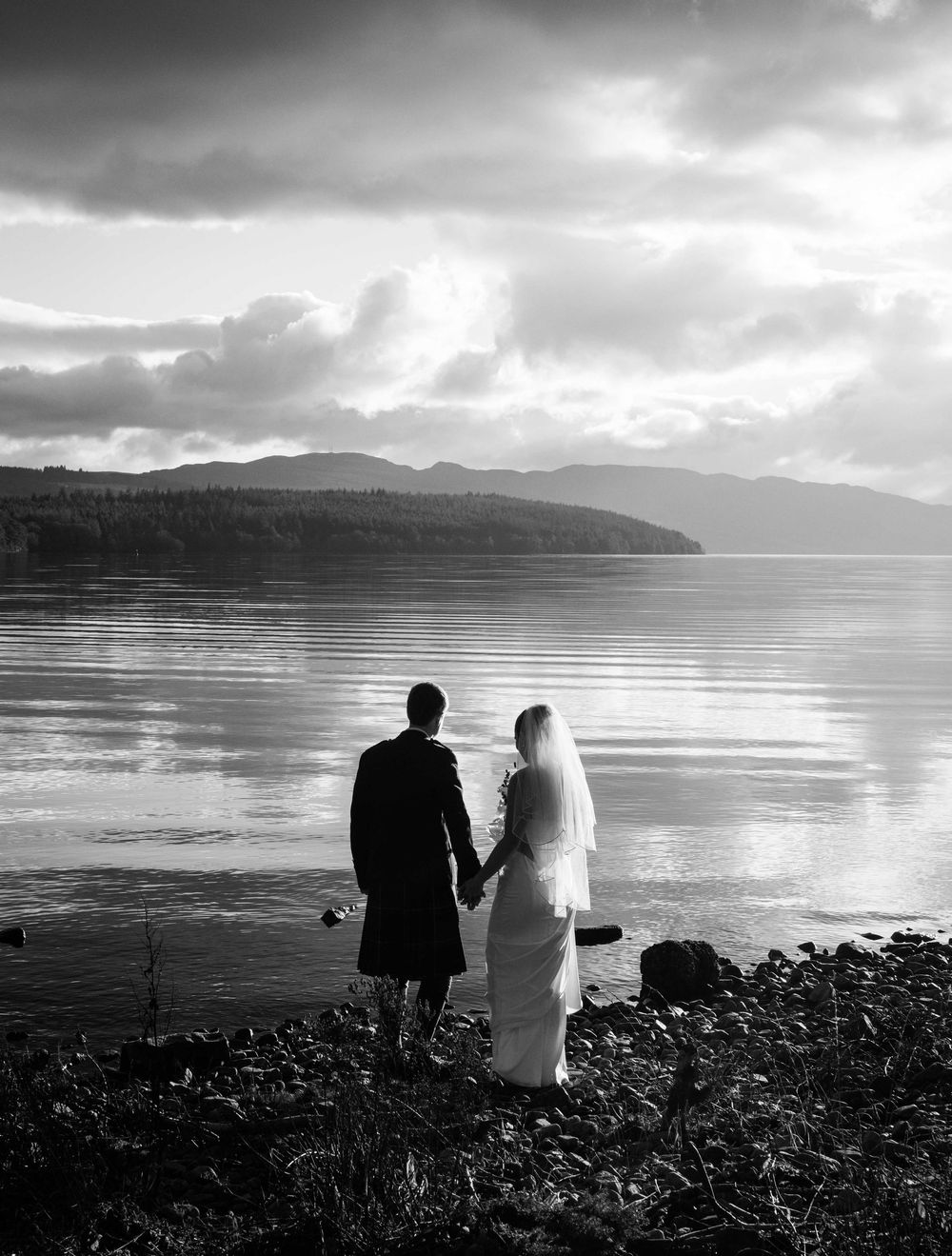 Black and white photo of a bride and groom holding hands by a serene lakeshore with mountains in the background.