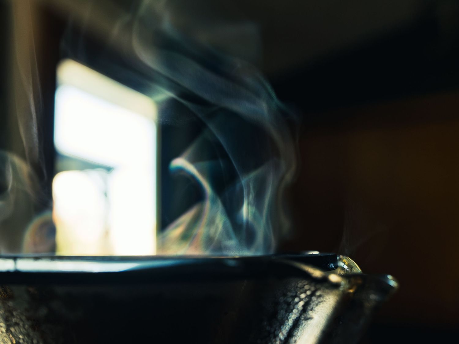 Wisps of steam curling off the rim of freshly brewed coffee, backlit by soft window light against a dark background.