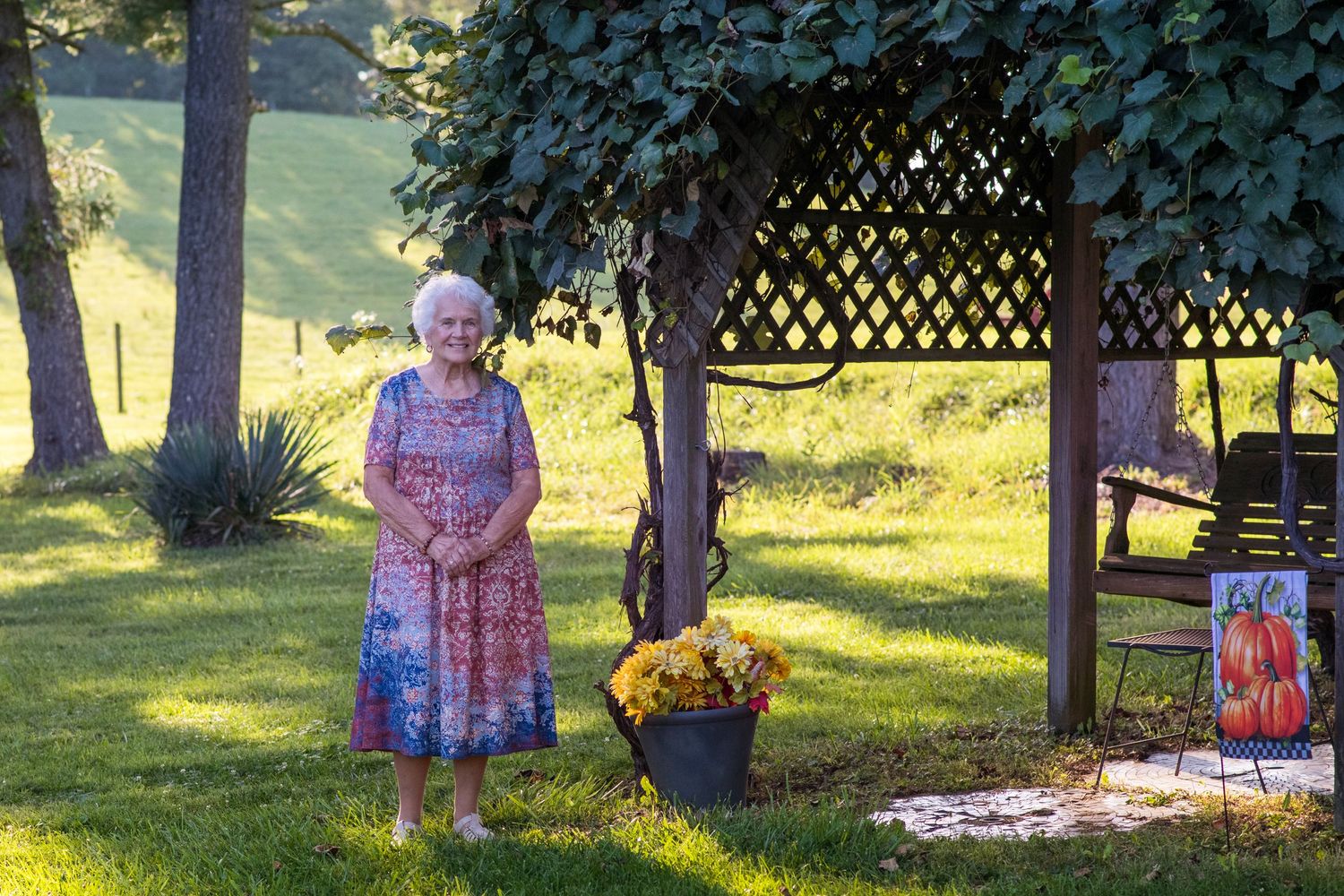 Elderly woman in floral dress standing by a garden pergola with yellow flowers and pumpkin decor on a sunny day.