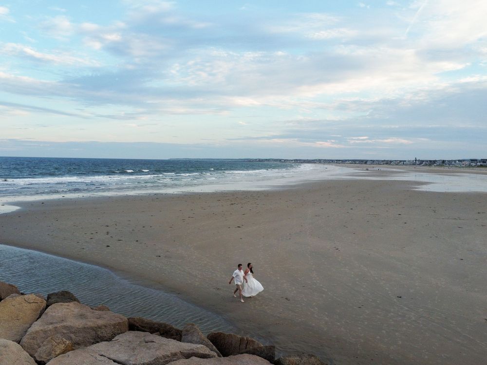 Aerial view of a bride and groom walking on a vast sandy beach near rocky shores with ocean waves in the background.