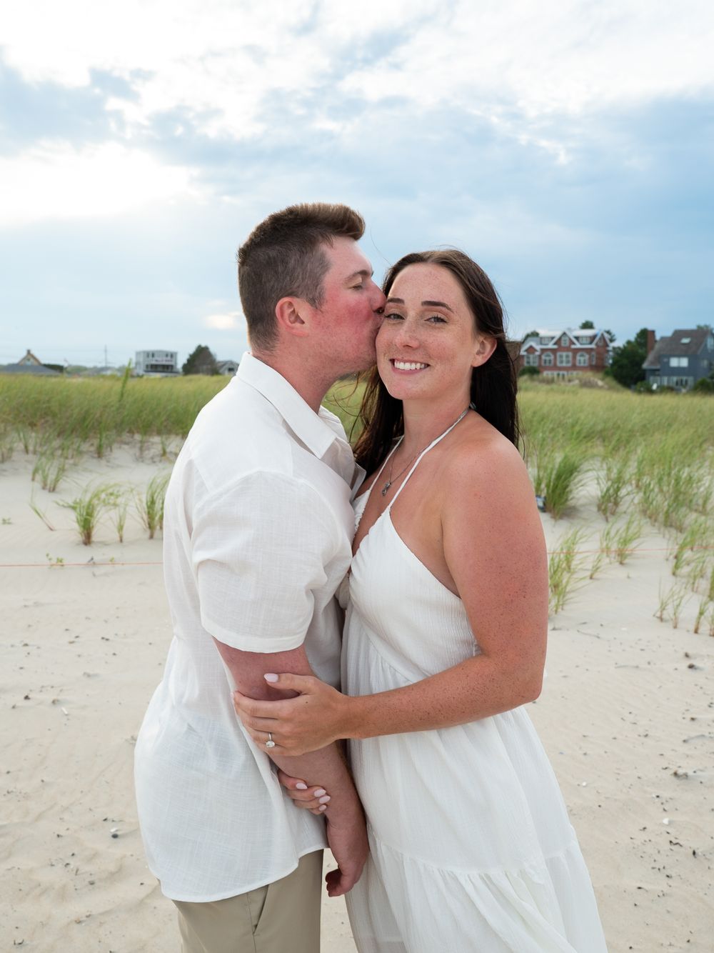Couple shares a romantic kiss on a sandy beach, woman in white dress smiling, grass dunes and houses in background.
