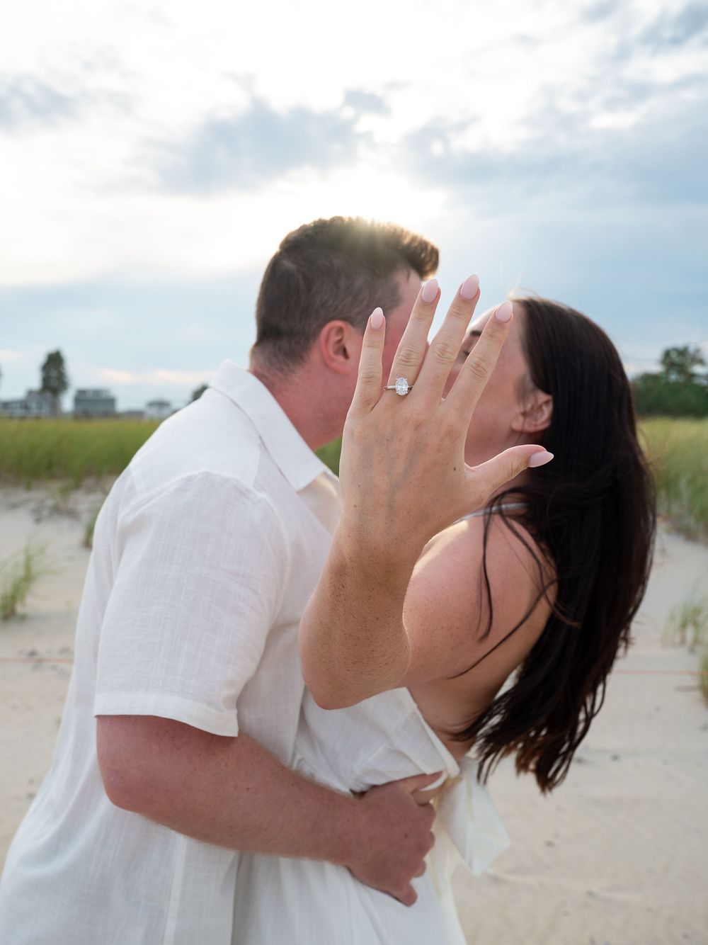 Couple kissing on beach while showing off engagement ring, sandy dunes and cloudy sky in background.