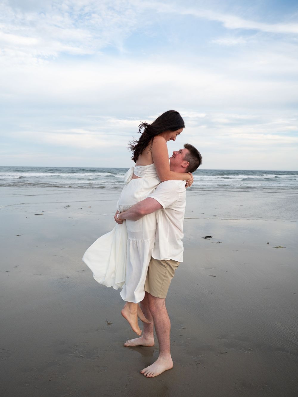 Couple embracing on a sandy beach, man lifting woman in white dress, ocean waves and cloudy sky in background.