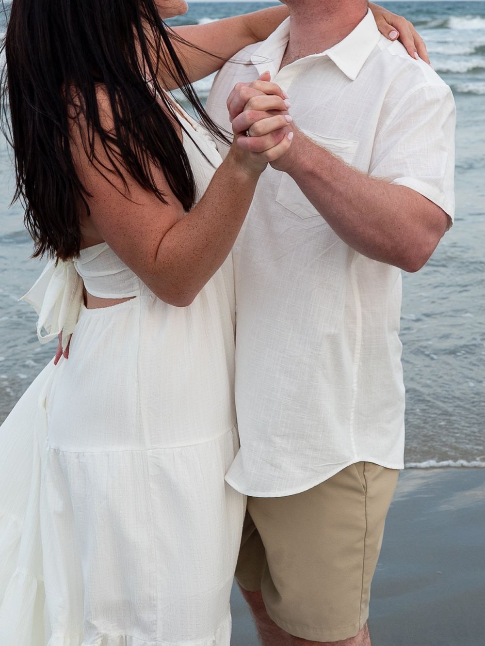 Couple dancing together on the beach, both dressed in white outfits, holding hands with ocean waves in the background.