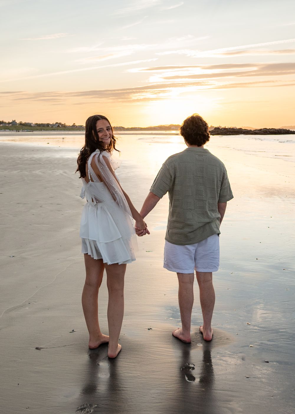 Couple holding hands on a sandy beach at sunset, woman in white dress looks back smiling, man in green shirt and white shorts.