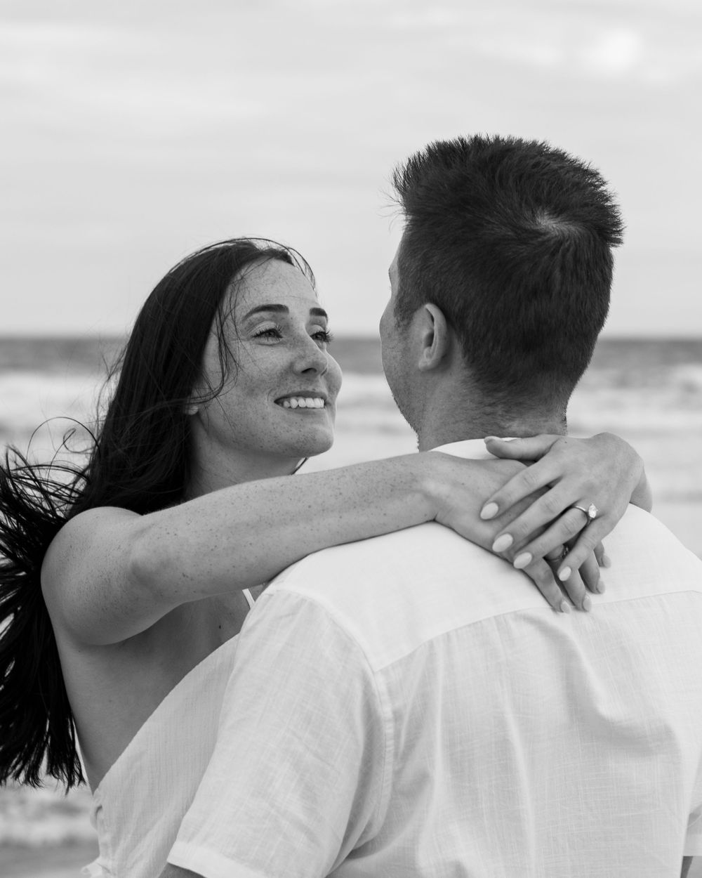 Black and white beach photo of smiling woman with dark hair embracing a man, ocean waves in background.
