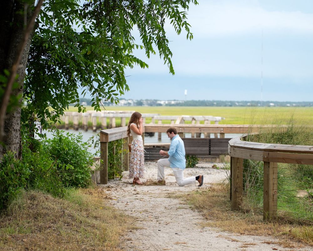 Man proposing on one knee to woman on a wooden dock overlooking a scenic marsh and waterway.