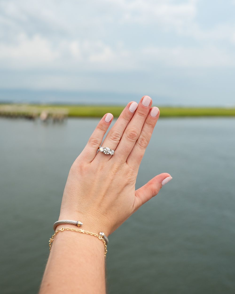 A hand with French manicure displaying a diamond engagement ring and gold bracelet, posed against a scenic waterway backdrop.