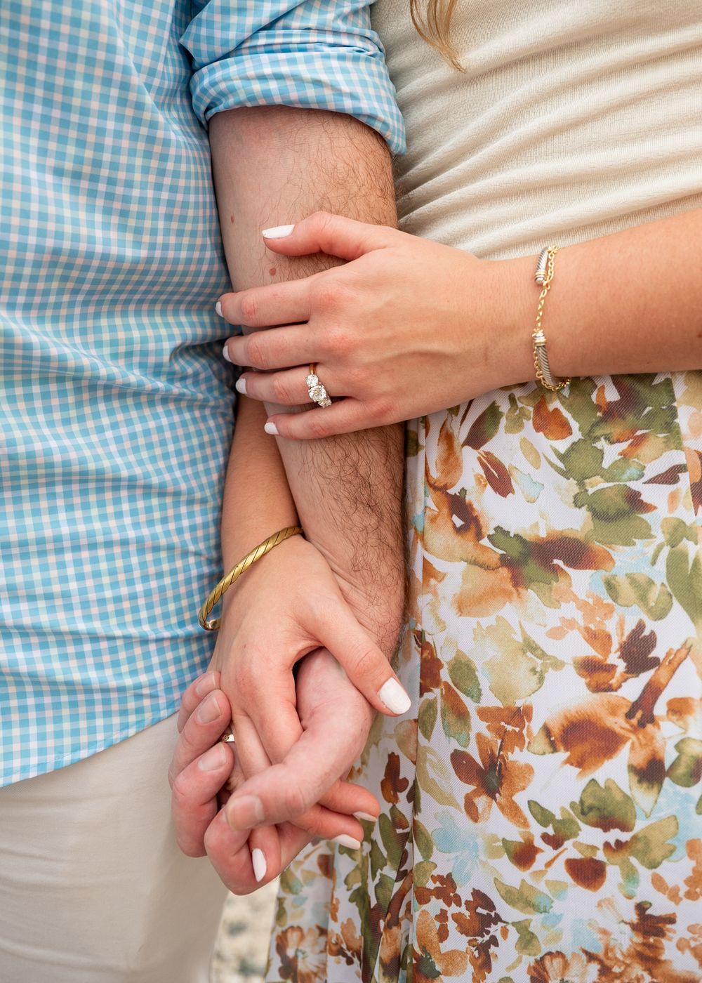 Couple holding hands showing engagement ring and gold bracelet, woman in floral dress, man in blue plaid shirt.
