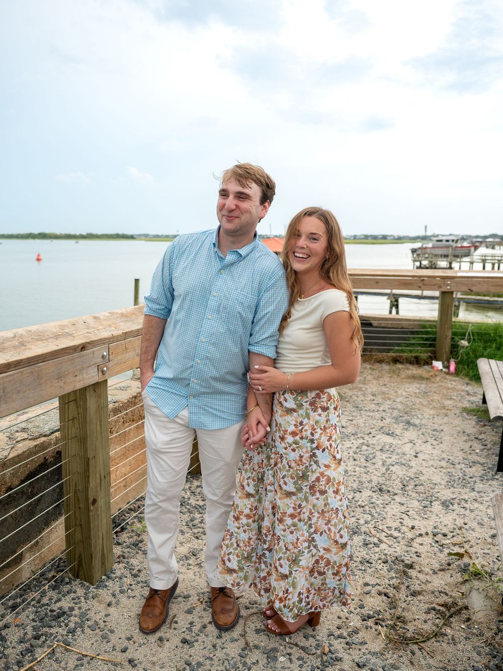 A smiling couple poses on a waterfront boardwalk, man in blue shirt and woman in floral skirt.