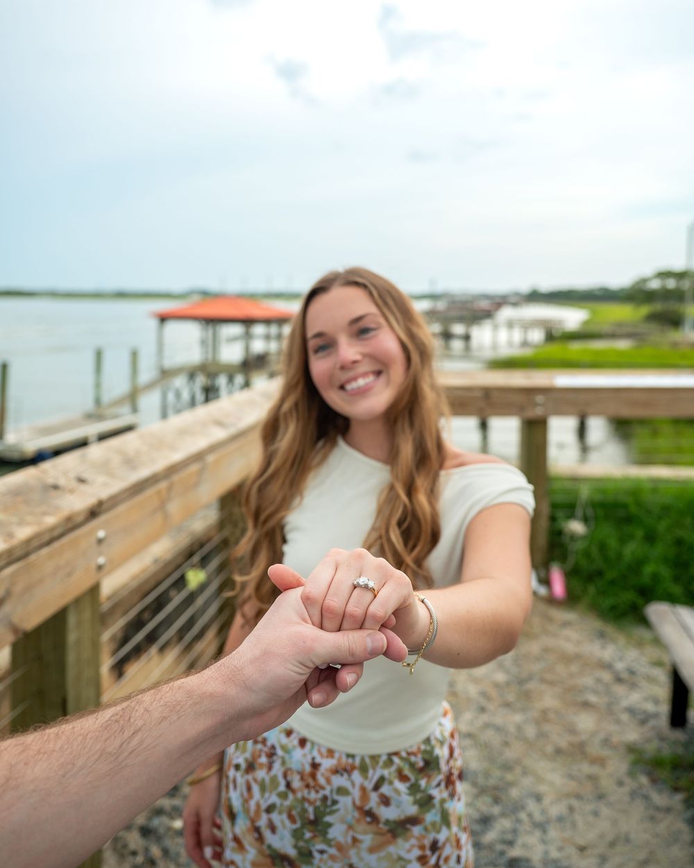 Smiling woman with long wavy hair shows off engagement ring near waterfront dock on cloudy day.