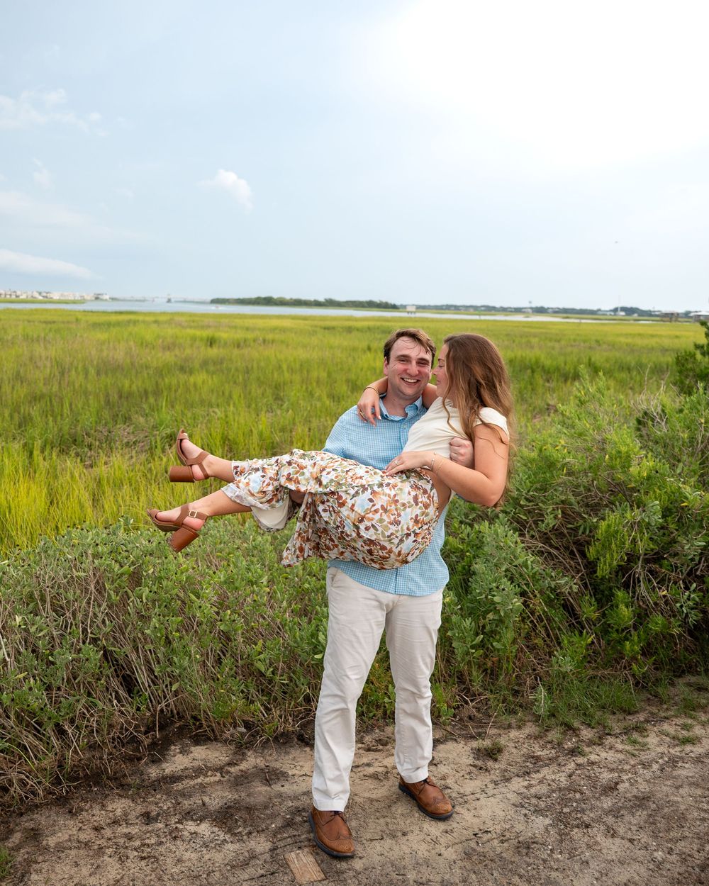 A man carries a woman in his arms while she kisses his cheek in a coastal marsh landscape.