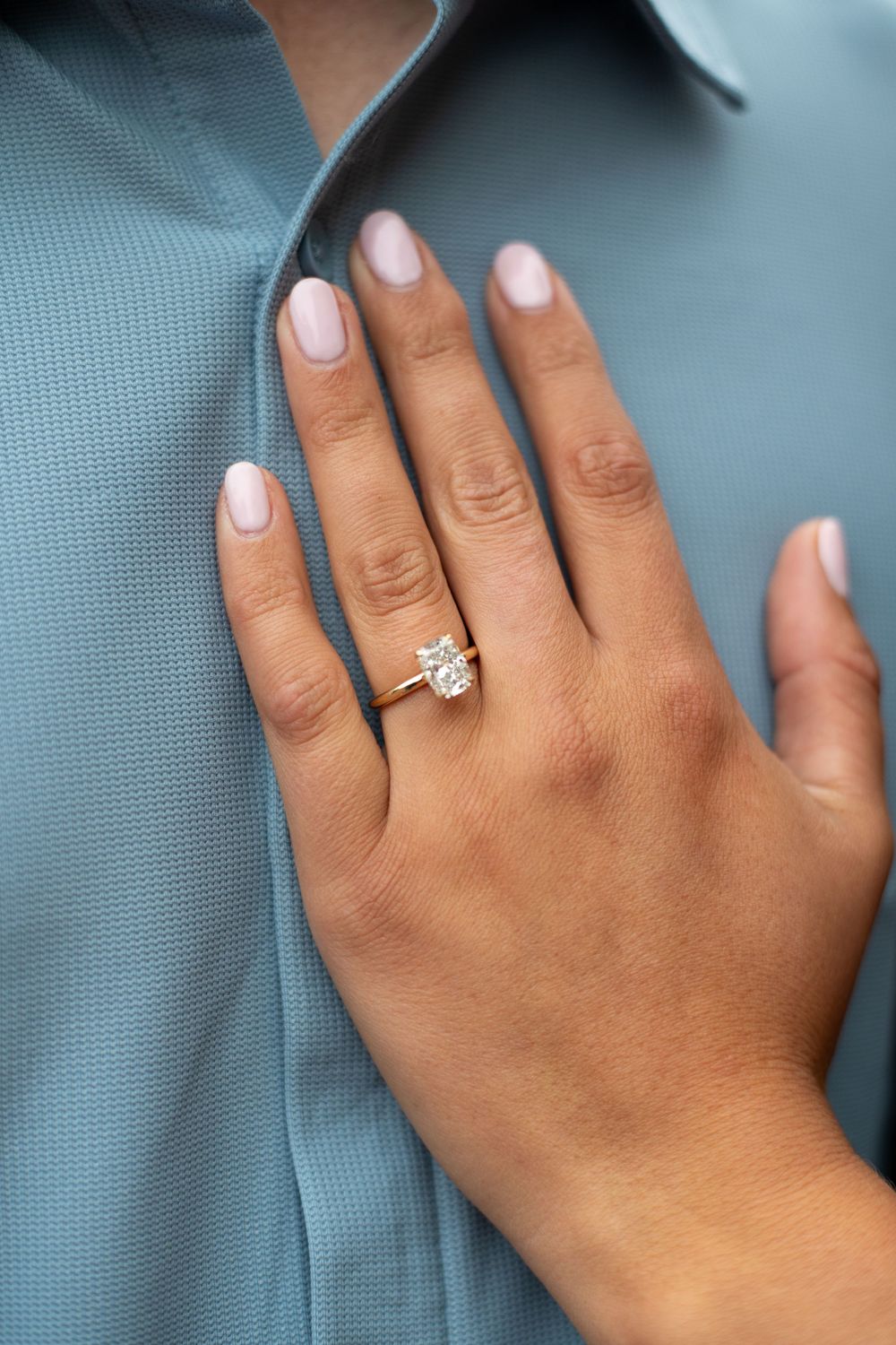 Close-up of a hand with pink nails wearing a gold solitaire diamond engagement ring on the ring finger.