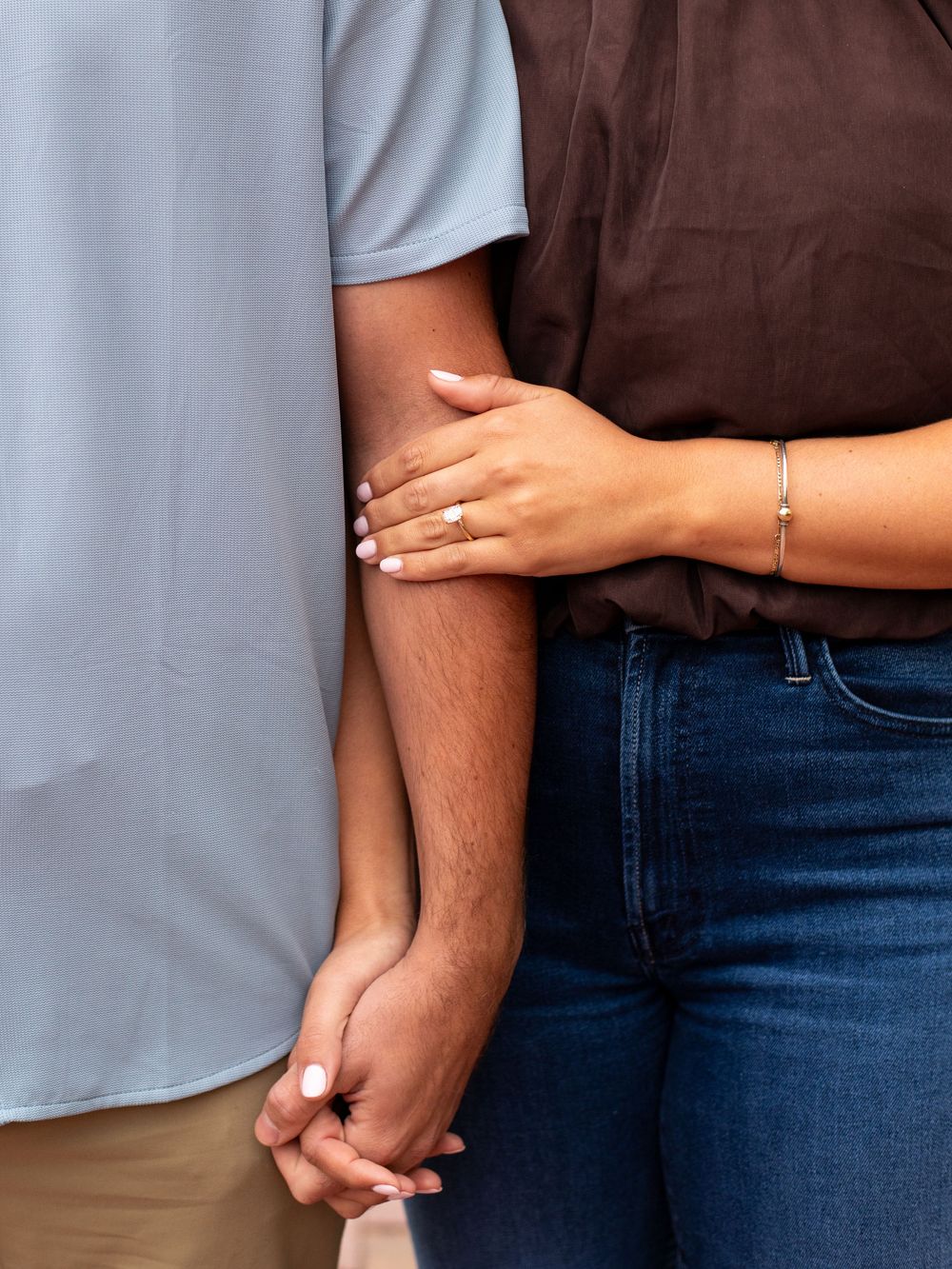 Couple holding hands, woman wearing engagement ring and bracelet, man in gray shirt, woman in dark jeans.