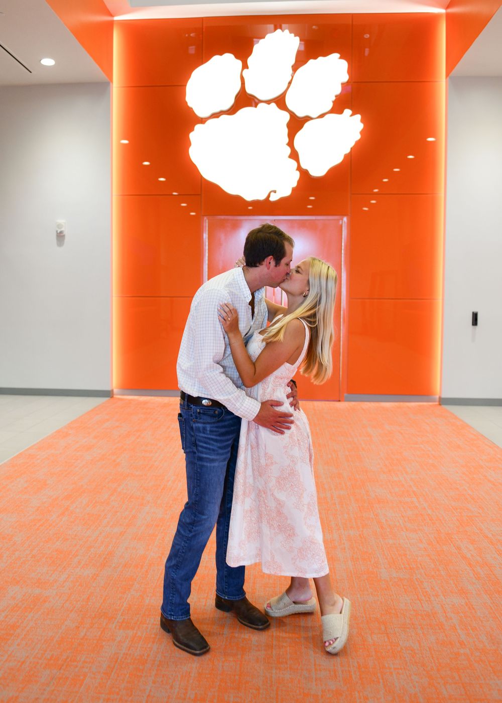 Couple sharing a romantic kiss in front of a Clemson Tigers paw logo on an orange wall.