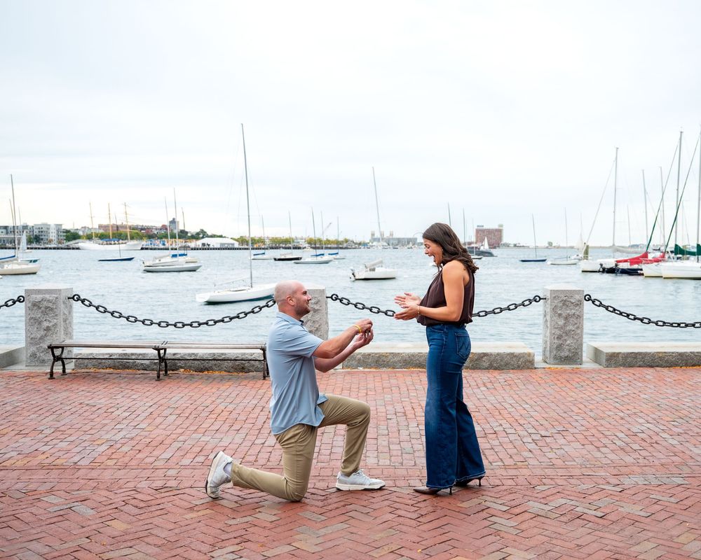 Man kneeling on brick waterfront proposing to woman with sailboats in harbor background.