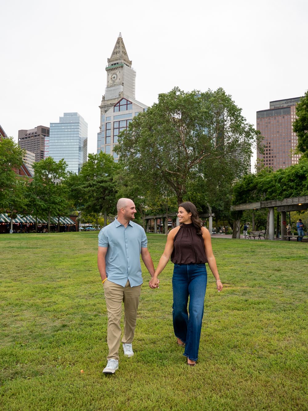 Couple holding hands walking through a green park with Boston city skyline and clock tower in background.