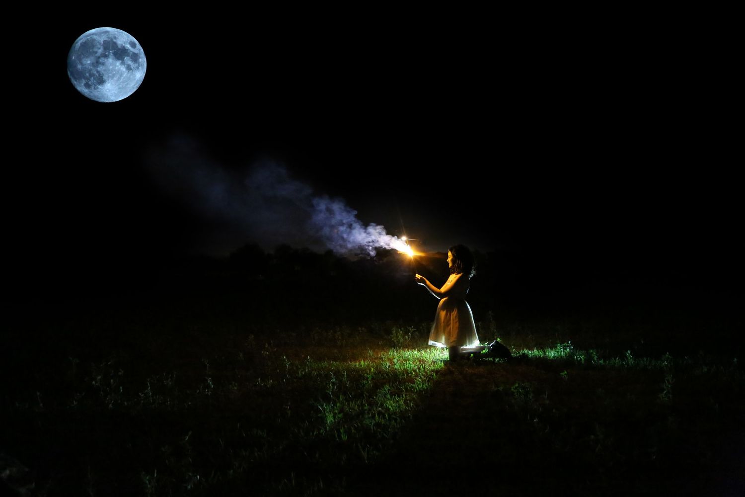 A person kneels in a dark grassy field holding a sparkler that trails smoke beneath a bright full moon.
