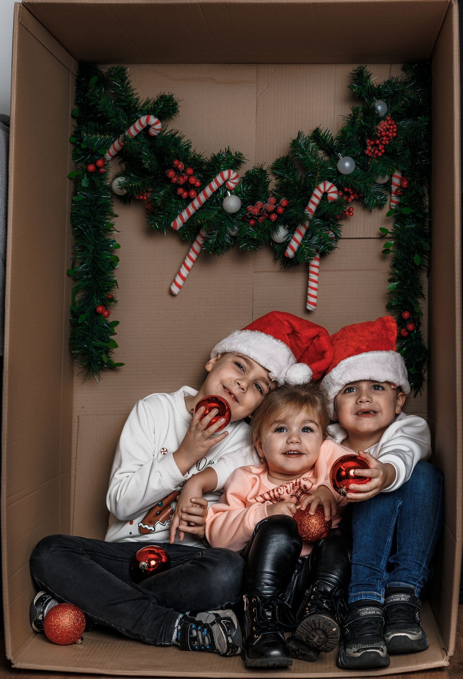 Three children in Santa hats sit inside a decorated cardboard box holding red Christmas ornaments.