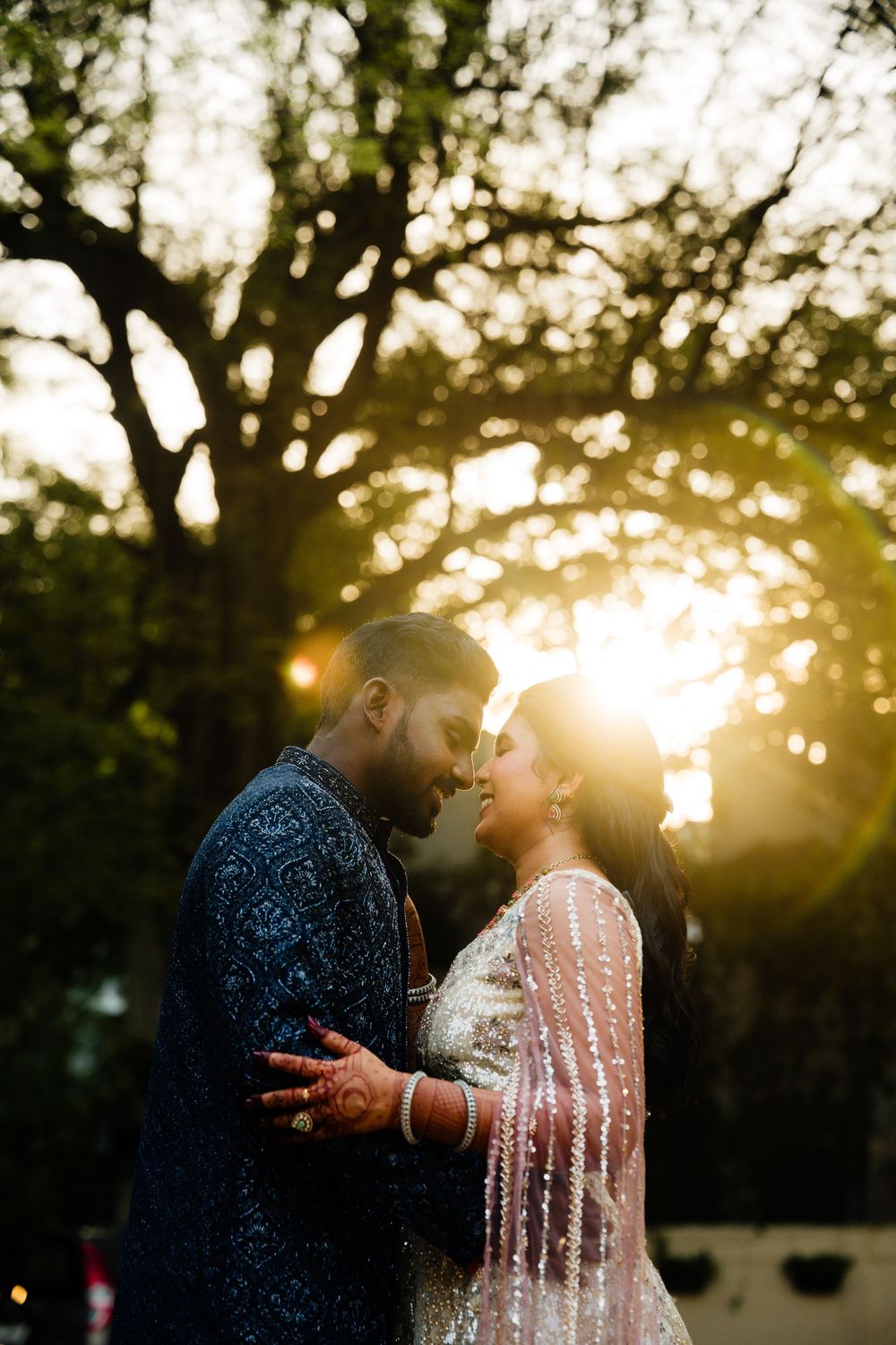 Indian couple in traditional wedding attire share a romantic moment at golden hour, backlit by sunset through trees.