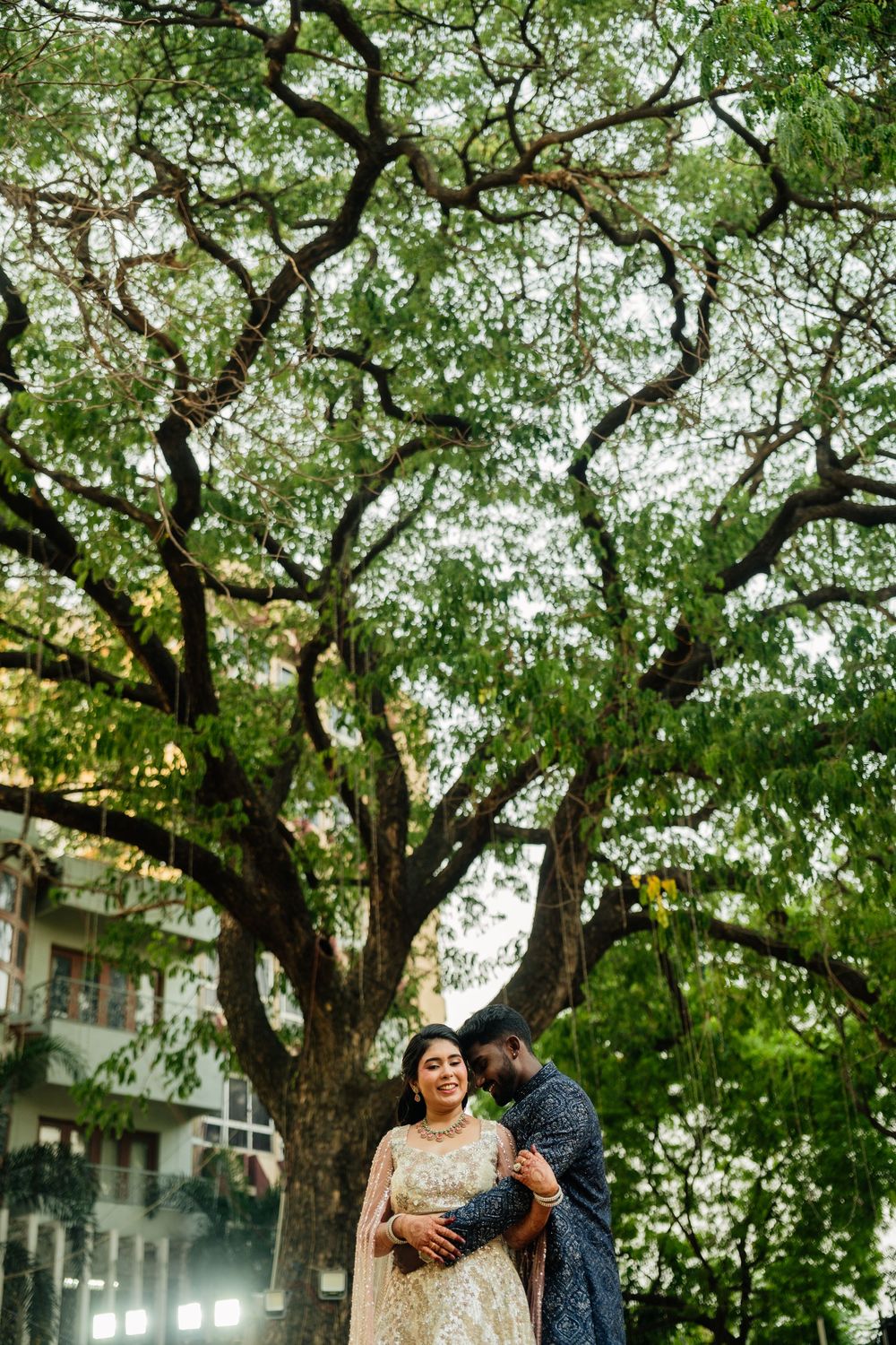 Couple poses romantically under a large tree, bride in embroidered lehenga, groom in dark outfit.