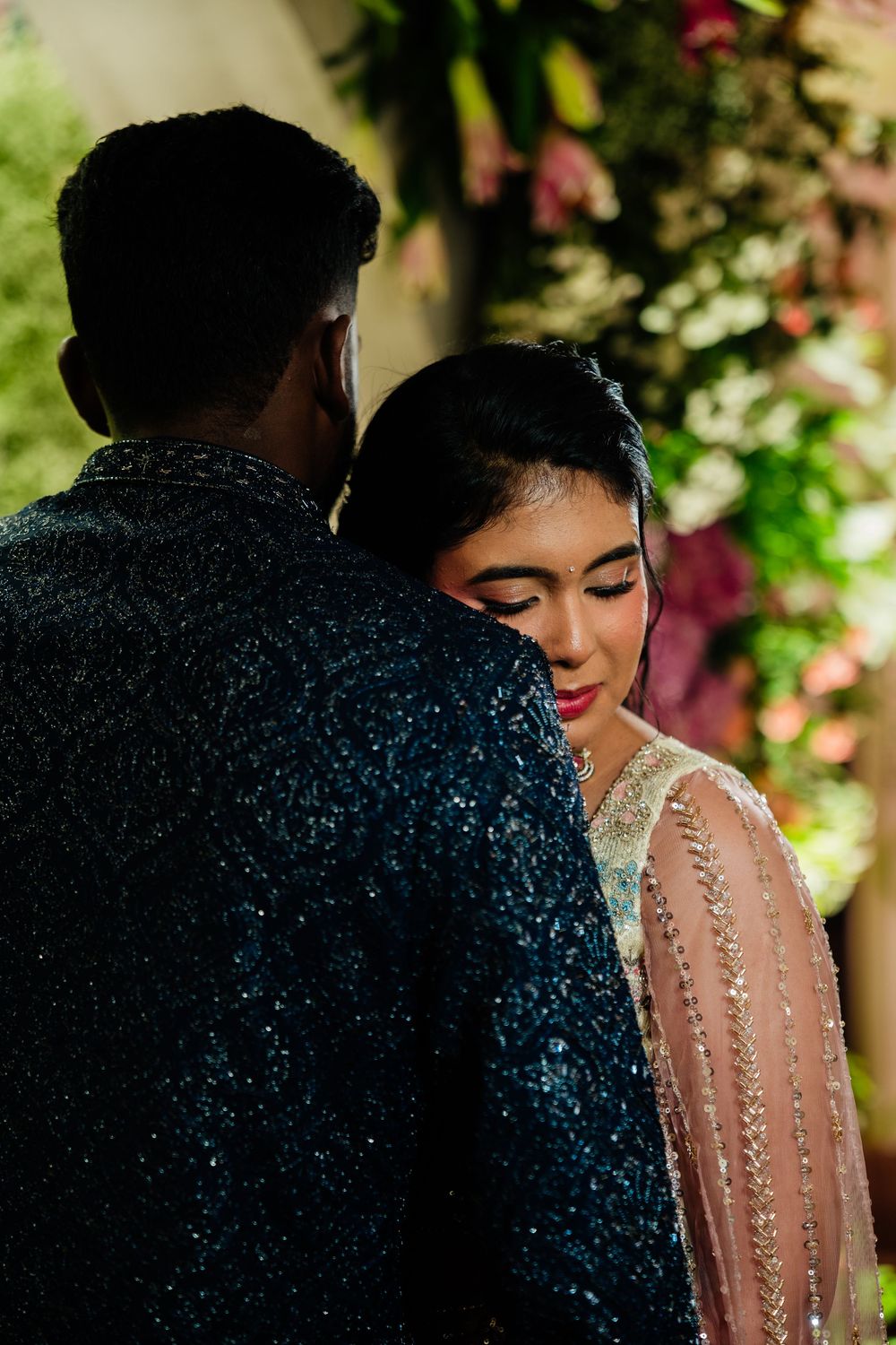 Bride in embellished outfit leans on groom in dark sequin sherwani at floral wedding backdrop.