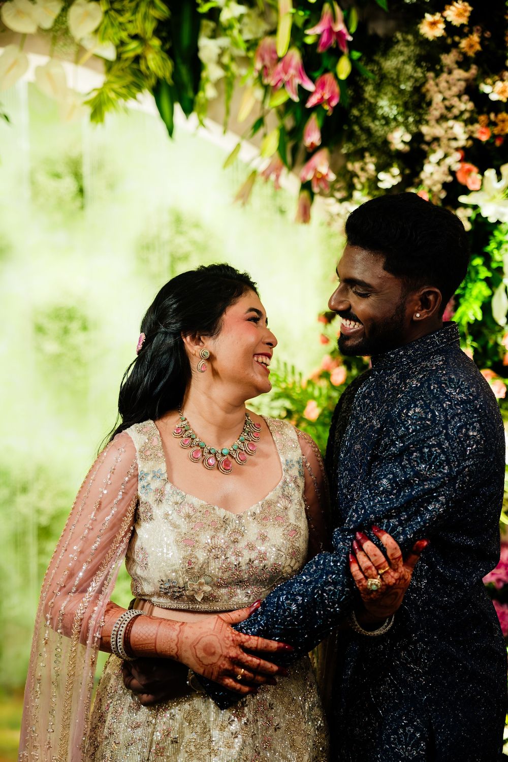 Joyful Indian couple in traditional attire laughing together at a floral wedding ceremony backdrop.