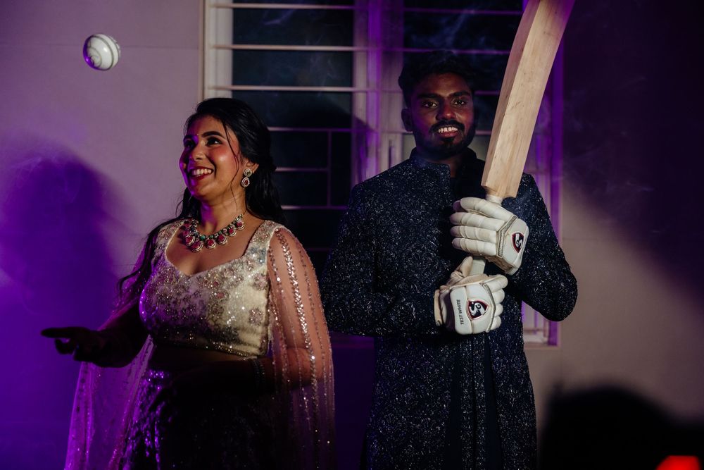 Indian couple in traditional attire, woman in embroidered lehenga and man holding cricket bat in batting gloves.