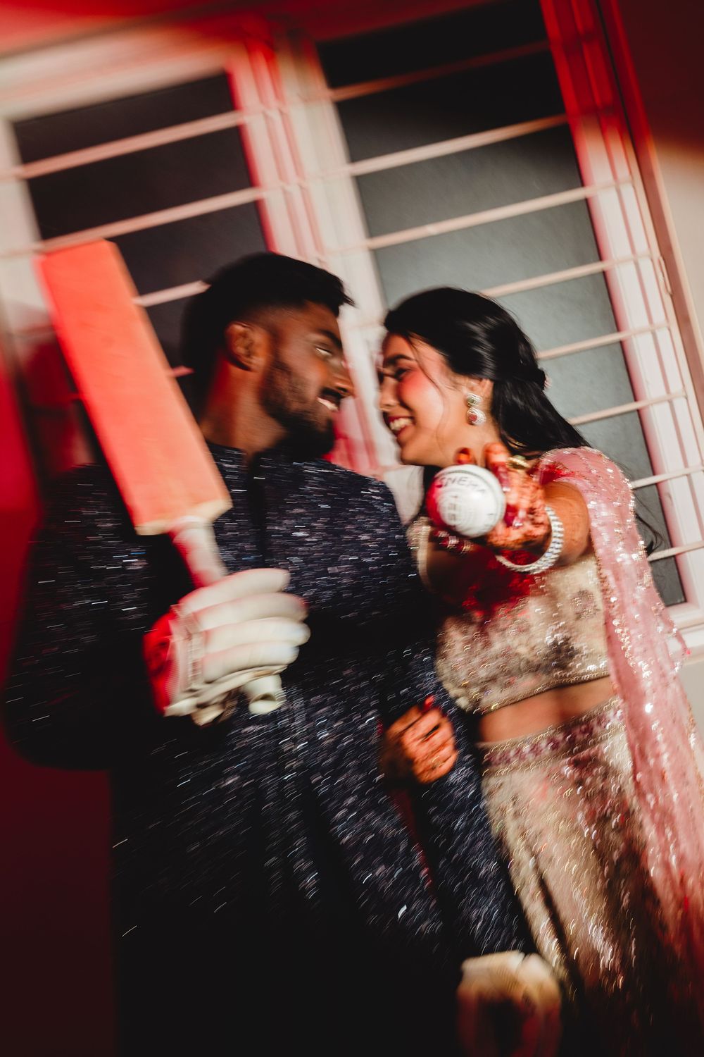 Indian bride and groom playfully pose with cricket bat and ball in traditional wedding attire during festive celebration.
