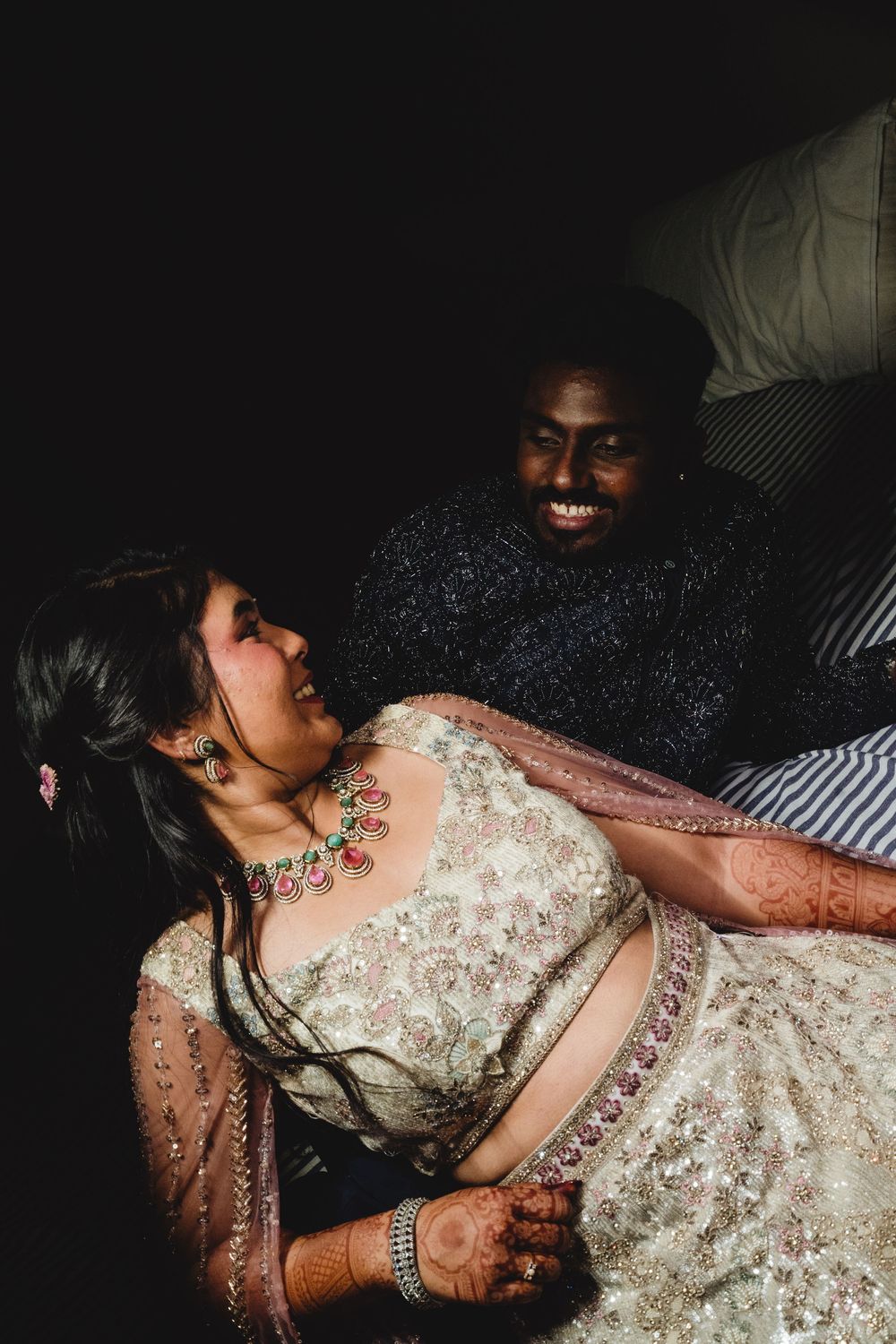 Couple laughs joyfully together, bride in embroidered lehenga with ruby necklace, groom in sequin outfit.