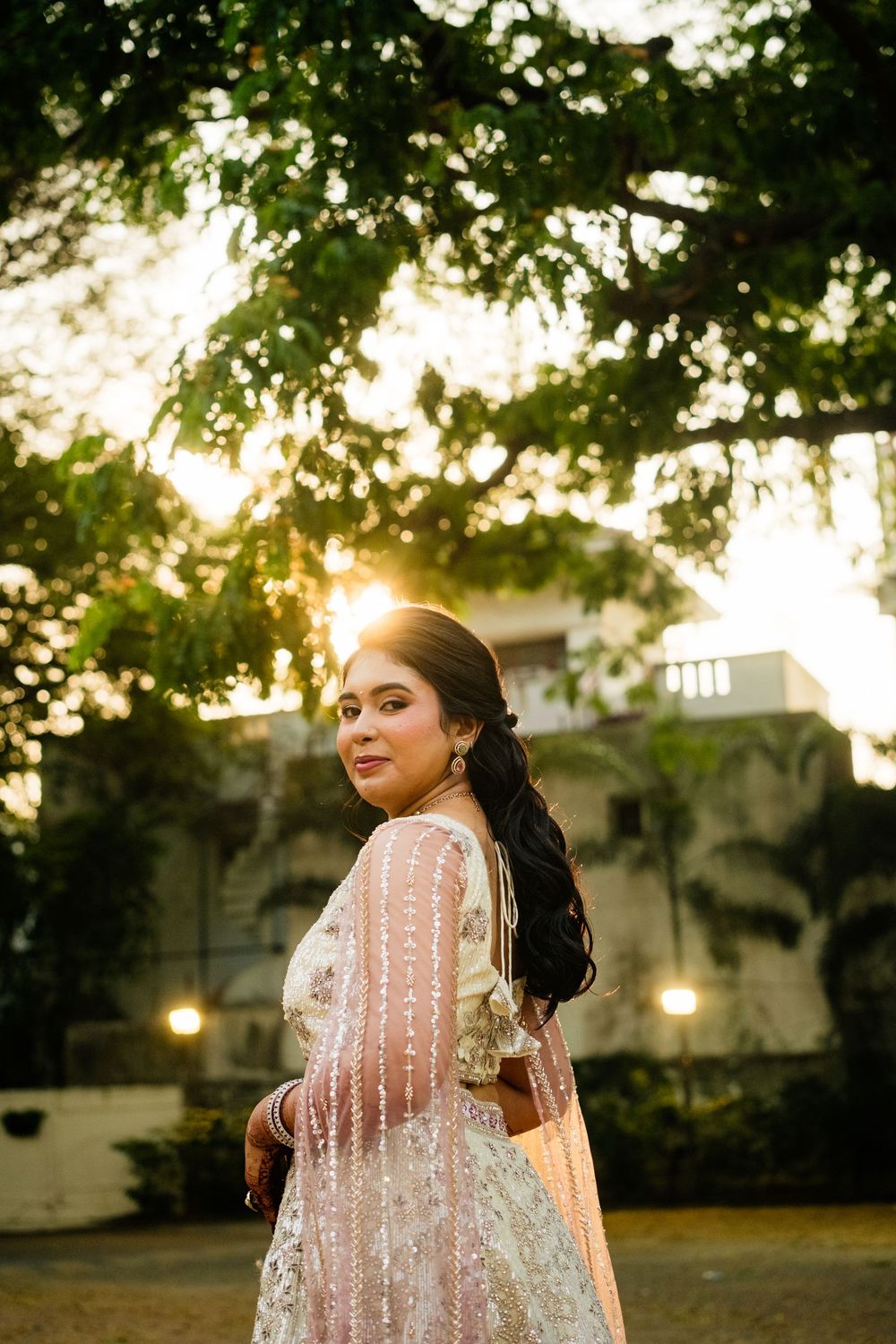 Bride poses gracefully at golden hour under lush trees in a white embroidered lehenga with dupatta.