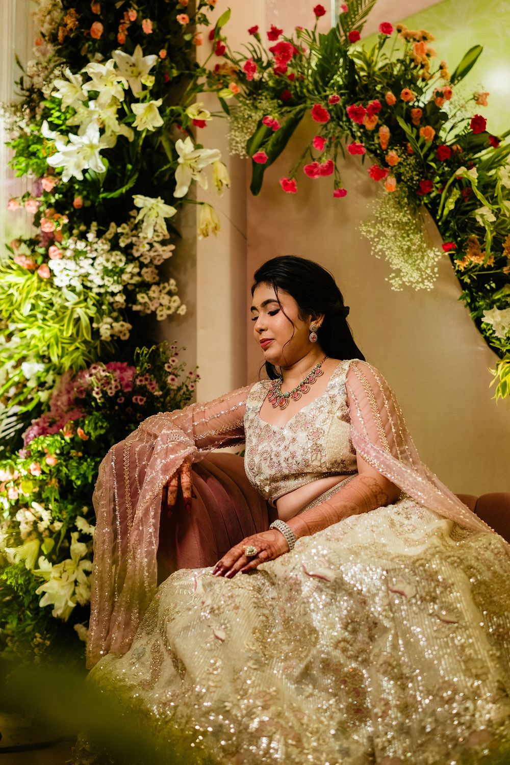 A bride in a shimmering ivory lehenga sits gracefully amid lush floral decorations with white lilies and red blooms.