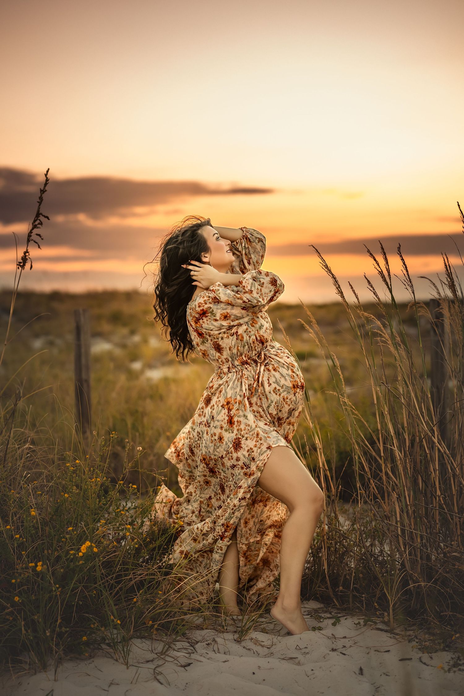 Woman in floral dress poses gracefully on sandy dunes at golden sunset, hair flowing in the wind.