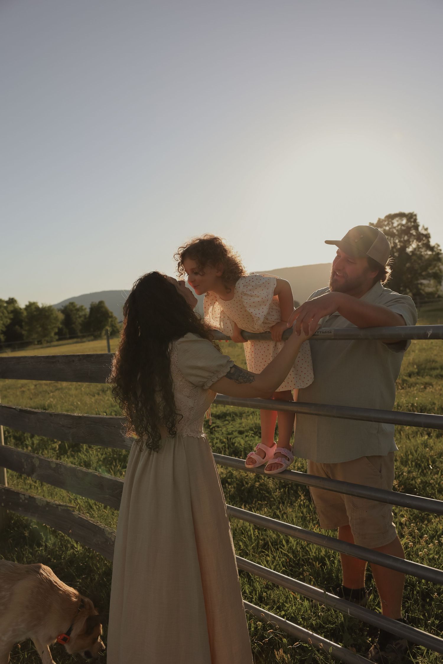 A family with a young child enjoys golden hour sunlight by a fence on a scenic green farm.