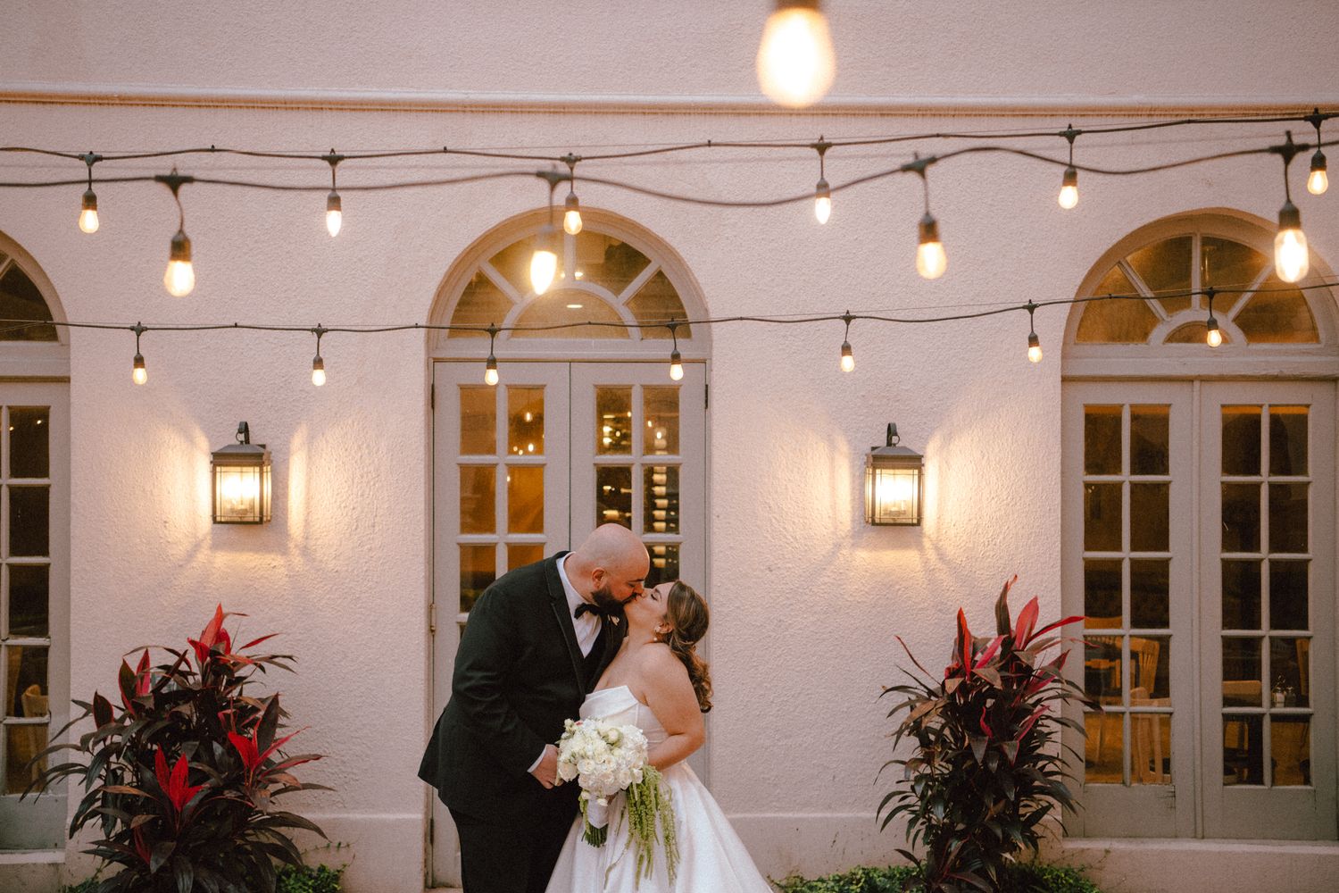 Bride and groom sharing a romantic kiss under string lights at an elegant outdoor wedding venue at night.