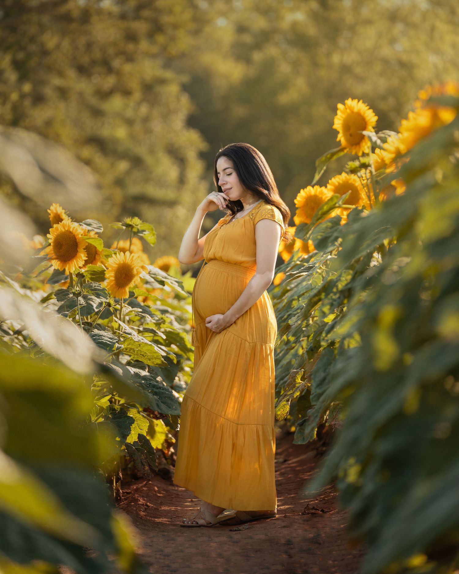 Pregnant woman in yellow dress standing among sunflowers in golden light, cradling her baby bump.
