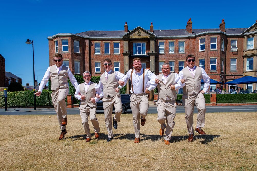 Seven groomsmen in matching tan suits with suspenders joyfully running outdoors on a sunny wedding day.