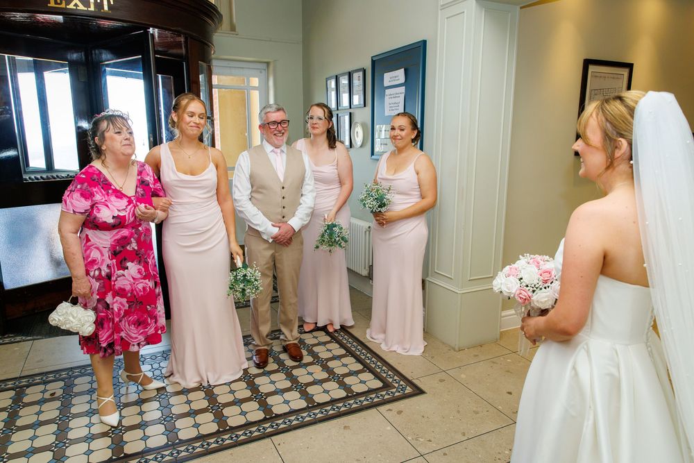 Bride in white gown holding bouquet faces bridesmaids in pink dresses with groom and family in elegant venue foyer.