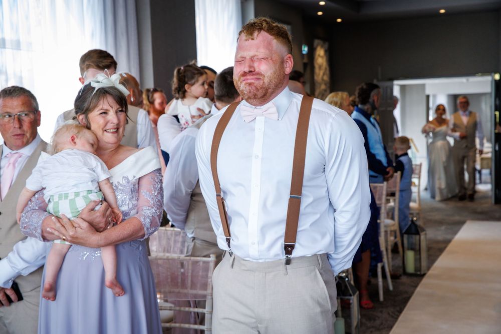 Man in white shirt and suspenders walks through wedding venue with guests seated in background.