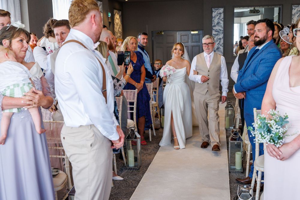 Bride walks down the aisle in elegant white gown at indoor wedding ceremony with guests in formal attire.