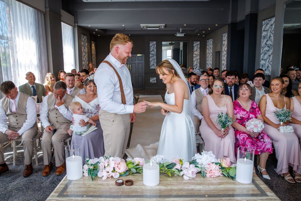 Bride and groom exchange vows at elegant indoor wedding ceremony with guests seated and floral arrangements on display.