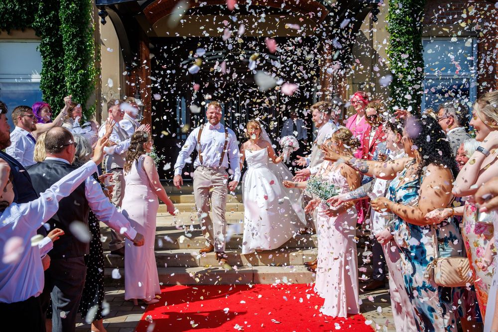 Newlyweds walk down red carpet through confetti shower surrounded by cheering wedding guests outdoors.