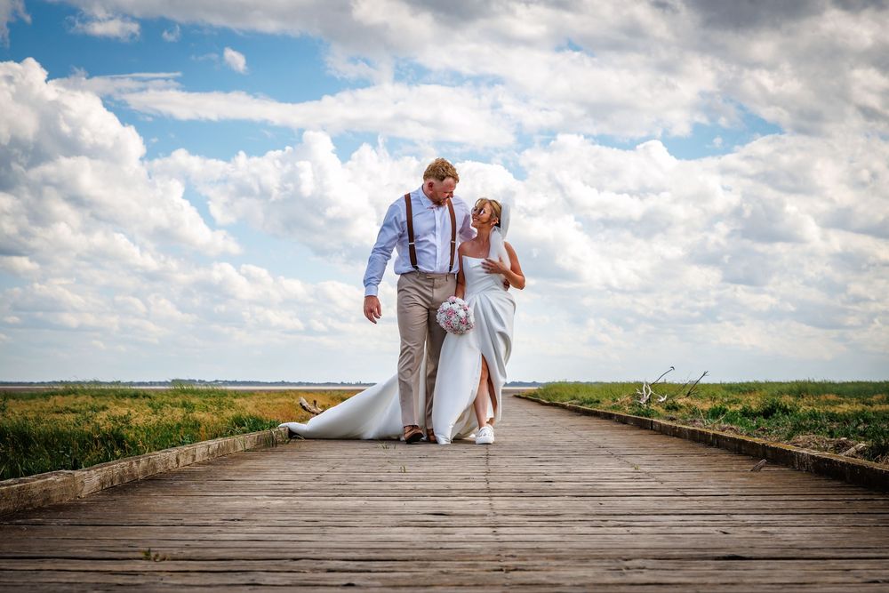 Bride and groom walking hand-in-hand on a wooden boardwalk under a blue sky with fluffy clouds on their wedding day.