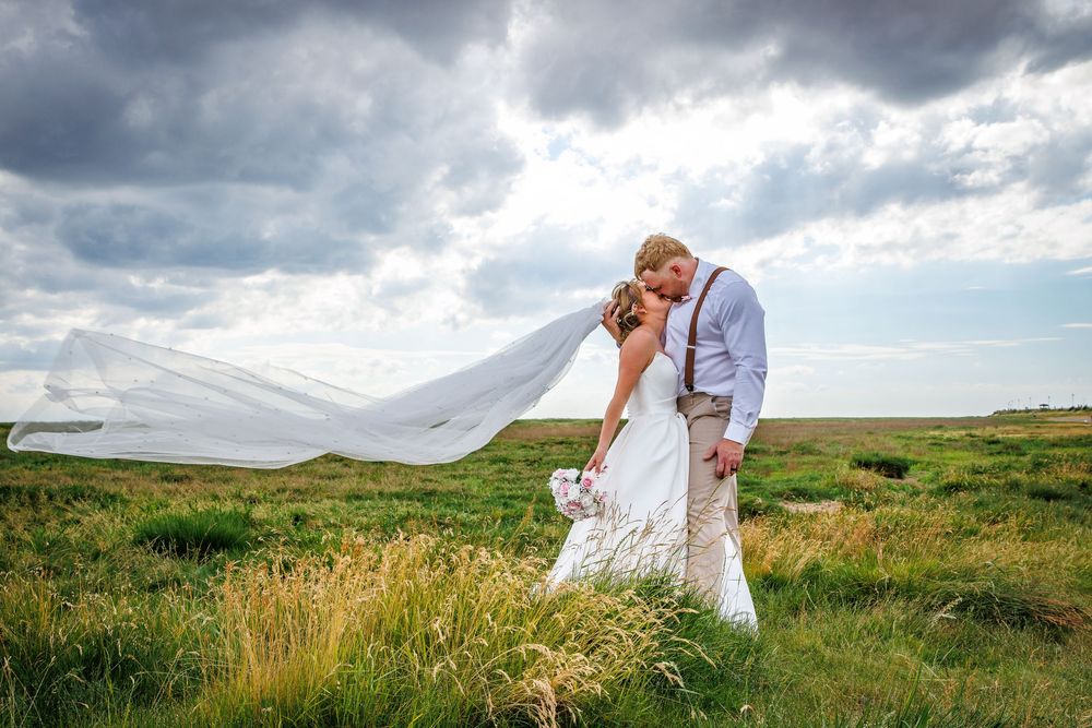 Bride and groom share a romantic kiss in a scenic field, with her veil flowing in the wind under dramatic cloudy skies.